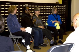 Picture shows from left to right, Project Manager Katherine Allen, Project Advisor Edery Herrera, Artist Teresa Jaynes, and Project Advisor Suzanne Erb, seated on grey folding chairs, in front of a card catalog. Allen, smiles and holds a sheet of paper in her lap. Hererra laughs, his left hand holding a tiny microphone near his mouth. Jaynes laughs wth her mouth open. Erb smiles and knits with a ball of yellow yarn under her right elbow. Parts of the backs of the heads and shouldPicture shows from left to right, Project Manager Katherine Allen, Project Advisor Edery Herrera, Artist Teresa Jaynes, and Project Advisor Suzanne Erb, seated on grey folding chairs, in front of a card catalog. Allen smiles and holds a sheet of paper in her lap. Hererra laughs, his left hand holding a tiny microphone near his mouth. Jaynes laughs with her mouth open. Erb smiles and knits a knitted square from a ball of yellow yarn tucked under her right elbow. Parts of the backs of the heads and shoulders of two audience members, separated by a chair, are visible in the foreground. [End of description]