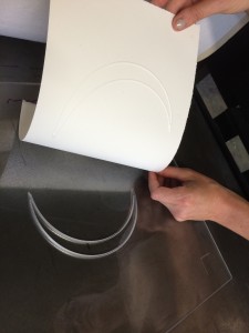 Crescent print and printing plate for Jaynes's The Moon Reader, 2014. Picture shows a translucent printing plate with the image of a crescent moon deeply etched into it. A white piece of paper with the crescent image embossed on it is being peeled away by the disembodied pair of hands of the printer. Her right hand, farthest from the viewer, holds the paper in the upper right corner. Her left hand, close to the viewer, is above the lower right edge of the bottom of the paper still attached to the plate. The reflection of the woman printer, who wears glasses, is seen in the lower edge of the printing plate, which is closest to the viewer. [End of description]