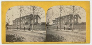 Picture shows two almost identical photographs on a cardboard mount. Photographs show the exterior of the multi-story, half block long brick building of the Pennsylvania Institution for the Instruction of the Blind. Building contains many rectangular-shaped windows. A tall picket fence lines the property. Three men and two women stand on the sidewalk evenly lined with trees in front of the fence. The men stand a few feet apart from each other and the women stand next to each other a few feet to the right of the men. The men wear dark-colored hats and suits. The women are attired in dark-colored hats and one is in a light-colored, long-sleeved dress with crinoline skirt, and the other in a dark-colored, long-sleeved dress with crinoline skirt. [End of description]