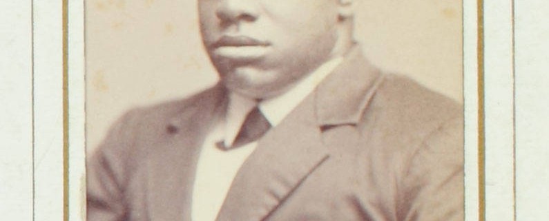 Thomas Greene Bethune, known as Blind Tom, ca. 1870. Black & white photograph. 4 x 2.5 in. Picture depicts the carte-de-visite portrait photograph of musician Thomas Greene Bethune, later Wiggins, known as Blind Tom. Shows the young African American man from his waist up, his body slightly angled to the viewer’s right. His tightly curled hair is shortly cropped. His eyes are closed. He wears a white shirt with a turned down collar. Under the collar is a dark cross tie. He also wears a dark jacket with wide notch lapels, several creases around the waist, and the top button fastened. The photograph is framed within a rectangular shape printed with a thick gold line surrounded by a thin black line. The frame is on light-colored paper. The top edge of the frame is slightly rounded. Hand written text below the portrait reads: “Blind Tom” [End of description]