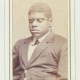 Thomas Greene Bethune, known as Blind Tom, ca. 1870. Black & white photograph. 4 x 2.5 in. Picture depicts the carte-de-visite portrait photograph of musician Thomas Greene Bethune, later Wiggins, known as Blind Tom. Shows the young African American man from his waist up, his body slightly angled to the viewer’s right. His tightly curled hair is shortly cropped. His eyes are closed. He wears a white shirt with a turned down collar. Under the collar is a dark cross tie. He also wears a dark jacket with wide notch lapels, several creases around the waist, and the top button fastened. The photograph is framed within a rectangular shape printed with a thick gold line surrounded by a thin black line. The frame is on light-colored paper. The top edge of the frame is slightly rounded. Hand written text below the portrait reads: “Blind Tom” [End of description]