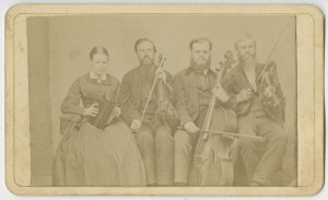 [Portrait of Hostetler family of blind musicians]. Mount Pleasant, Penna.: From A. N. Staufer, [ca. 1866]. Black & white photograph. 4 x 2.5 in. Picture shows one woman and three men, seated next to each other, and holding instruments. The woman holds an accordion in her lap and she looks slightly down. To her left is a man, his eyes closed, who holds a viola perpendicular to his lap with one hand and a bow in his other. To his left is a man resting a cello between his legs. He holds a bow across the base of the cello with his right hand. To his left is the last man, his eyes closed, who holds a violin by his left shoulder and a raised bow in in his right hand. The woman, as well as the man who holds a cello, wear glasses. The woman wears a dark-colored corseted dress with long sleeves and a long skirt. The men, who look toward the viewer, are bearded and wear dark-colored suits. [End of description]]