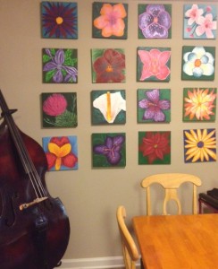Picture shows the tan painted wall of a dining area decorated with a display of twelve flower paintings. Paintings are arranged in vertical and horizontal rows of four. A cello rests near the paintings in the lower left corner of the image. In the right, toward the viewer, is the upper corner of a rectangular light-colored wooden table. A light-colored wooden chair is placed at the head and the side of the table. [end of description]