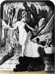 Picture depicts a composition inspired by folk-art devotional paintings known as retablo. Picture shows two nude female models, painted white to resemble sculpture, and staged on a pedestal covered in flowing drapery. To the viewer’s left stands a woman, her light colored hair pulled back into a low bun, her shoulders slightly pulled back and her arms stretched out from her sides. Her right leg is bent back and up toward her buttocks. She holds an upright sword in her right hand. She gazes down, to the viewer’s right, at a seated nude female figure with shortened arms and legs. Her back is to the viewer. Her dark wavy hair is pulled back into a low bun, and her left arm reaches to the standing figure. Her right arm rests on a small table covered with a white cloth. A vase of flowers adorns the table. The figures are in front of an elaborate backdrop. In the far left is a large nude photograph of the forward standing model. In the photo, she is standing with her left arm curved above her head and her right arm bowed down past her waist. Her right hand touches her inner left thigh. In the far right, next to the photograph, is a painted, shadowed, and winged form looking at a hand of salvation. A skeleton model lounges on the far left side of the scene. He is partially covered in drapery. He looks toward the women. The flooring extending from the front to the back of the scene is decorated with fleur-de-lis like details. Three lines of writing – the Retablo prayer – adorn the lower edge of the photograph on an attached yellow strip of paper. [end of description]