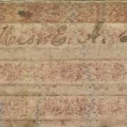Picture shows the upper edge of a writing board or tablet over a white background. The tablet is made of a brown cardboard-like material with a faded pink and blue marbled pattern and has raised, tactile, evenly spaced bars on its surface. In the center of the first bar, handwritten script reads “Mrs. E. A. Lusk.” [End of description]