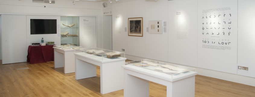 Picture shows exhibition installation shot of Touching the Book: Embossed LIterature for Blind People, taken from the back of the room and facing the exhibition entrance at the further end of the room on the right hand side. The flooring is light wood, and the walls are painted white. The photograph shows three free-standing display cases all containing 4-5 books opened and resting on cradles; two framed items on the right hand side of the wall as well as a large vinyl graphic of Moon's alphabetic system; and information panels are installed on the wall. At the further end of the room, and to the left of the entrance, is a large fixed display cabinet with three shelves holding artifacts (books and a writing frame). To the left of the cabinet is a blank TV screen and below that a table with a red covering that has been laid out with glasses of wine. [end of description]