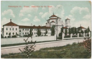 Picture shows an elongated white multi-story building with a central tower and columns. Building contains red pitched roofs and several rectangular and curved-edged windows. Trees evenly line the property in front of the building. A fence made of white stone wall on the left, an open gate in the center, and metal pickets on the right also lines the property in front of the trees. Clouds hang in the sky above the building. A grass lawn and bushes with pink flowers are visible across the street from the school and in the foreground. Red text printed in the upper right corner reads: “Philadelphia, Pa. Penna. Institution for the Blind.” [End of description].