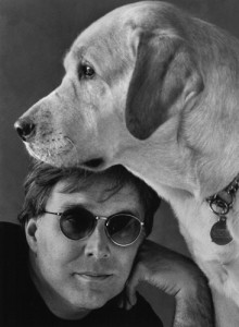 Picture shows bust-length portrait of Stephen Kuusisto with his dog. In the lower left edge of the image to the viewer, Kuusisto rests his left cheek on the knuckles of his left hand. He wears dark-shaded glasses and a black T-shirt. Above his head is the head of a yellow labrador posed in left profile. The dog wears a metal choke collar with a dog tag.