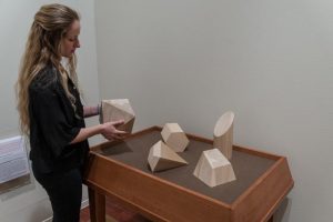 Picture shows a woman, in the far left, holding one of five differently-shaped wooden geometric forms that rest on a wooden table. The top is covered in brown linen. Shapes include a trapezoid, trapezium, octagon, slant and flat-edged cylinder, and prism. The woman wears a black shirt and pants, and has her long blond hair pulled slightly away from her face.