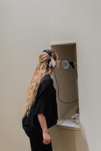 Picture shows a woman standing in front of a niche in a wall. She wears a headset, its cord extending from within the nice. Her face is near a funnel at the end of a mic-like stand. She wears a black shirt and pants, and has her long blond hair pulled slightly away from her face.