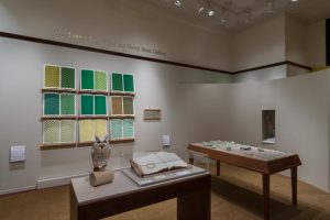 Picture shows the gallery, including four of Jaynes’s seven installations on view. In the center foreground is Jaynes’s Gift#1 on display on a flat, horizontally-angled rectangular table with a dark wood base. A white and brown paper owl rests on a cylindrical stand to the left of a large open book under a rectangular, clear acrylic hood. To the right of the display with the owl stands Jayne’s Gift #5, a map representation of the travels the 19th–century blind surveyor John Metcalf. It is a vertically-positioned, large rectangular table composed of a a linen top and a light brown wood base. A multi-color grid, outlines of geometric shapes, and green porcelain geometric shapes adorn the linen top. In the center background, on the back wall painted off-white, is Jayne’s Gift #4, a visual transmutation after the musical work of blind African American musician Thomas Wiggins. It is three horizontal and three vertical rows of prints in a geometric interplay of greens, browns and yellows. To the right of the prints on the wall is a small wooden frame in which brass musical notes are displayed. To the far right background is a view of Jaynes’s Gift #6, the scent mechanism the olfactometer, in a niche in an off-white curved wall. The floor of the room is covered with a tan colored carpet.