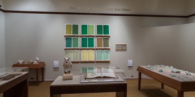 Picture shows the majority of the gallery, including four of Jaynes’s seven installations on view. In the center foreground is Jaynes’s Gift#1 on display on a flat, horizantally-angled rectangular table with a dark wood base. A white and brown paper maiche owl rests on a cylindrical stand to the left of a large open book under a rectangular, clear acrylic hood. To the right of the display with the owl stands Jayne’s Gift #5, a map represention of the travels of the 19th–century blind surveyor John Metcalf. It is a vertically-positioned, large rectangular table composed of a a padded linen top and a light brown wood base. A multi-color grid, outlines of geometric shapes, and green porcelain geometric forms adorn the linen top. To the left of the owl display is a partial view of a case of historical materials. In the left backround is Jayne’s Gift #3 inspired by the mathematical tools of the blind mathematician Nicholas Saunderson. It is a light brown table on which several large-sized wooden geometric shapes of different styles rest. In the center background, on the back wall painted off-white, is Jayne’s Gift #4, a visual transmutation after the musical work of blind African Amerian musician Thomas Wiggins. It is three horizontal and three vertical rows of prints in a geometric interplay of greens, browns and yellows. To the right of the prints on the wall is a small wooden frame in which brass musical notes are displayed. To the far right background is a partial view of an off-white curved wall. The floor of the room is covered with a tan colored carpet.