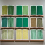 Picture shows the installation, which consists of nine screen prints across facing pages. They are displayed on wall mounted wooden shelves, spaced apart, in 3 vertical and 3 horizontal rows. The prints embody a visual translation of Wiggins’s music in a geometric interplay of greens, browns and yellows. Partially visible to the left is a wooden frame containing brass musical notes.