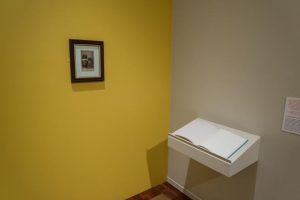 Picture shows a yellow painted wall on which only a wooden frame with a late 19th-century black and white cabinet card portrait hangs in the center. The portrait shows a couple. To the lower right is an open book of white pages, Jayne’s Gift #7, resting on a pedestal that juts from an off-white painted wall.