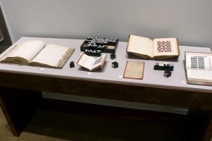 Picture shows the top of a case displaying (left to right) an open book of raised print; mathematical blocks with the upper case roman letters T,V, and L; a book open to a square-shaped illustration; a scrapbook open to a page with a paper pattern composed of a pink background with repeated rows of maroon colored crosses interspersed with rows of maroon colored squares; a sepia-toned sheet of paper on a red background; mathematical blocks; and a book resting vertically and opened to a grid-shaped illustration across from a page of printed text. The materials lay on grey linen.