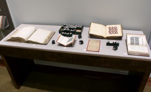 Picture shows the top of a case displaying (left to right) an open book of raised print; mathematical blocks with the upper case roman letters T,V, and L; a book open to a square-shaped illustration; a scrapbook open to a page with a paper pattern composed of a pink background with repeated rows of maroon colored crosses interspersed with rows of maroon colored squares; a sepia-toned sheet of paper on a red background; mathematical blocks; and a book resting vertically and opened to a grid-shaped illustration across from a page of printed text. The materials lay on grey linen.