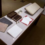 Picture shows the top of a case displaying (left to right) two grooved writing boards in pink and black hues; two letters in manuscript; a box of small blocks with its slatted cover removed and beneath it; a reproduction of an illustration of Victorian writing frames; and an open book with white pages of raised print. The historical materials lie on grey linen.