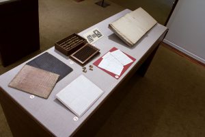Picture shows the top of a case displaying (left to right) two grooved writing boards in pink and black hues; two letters in manuscript; a box of small blocks with its slatted cover removed and beneath it; a reproduction of an illustration of Victorian writing frames; and an open book with white pages of raised print. The historical materials lie on grey linen.