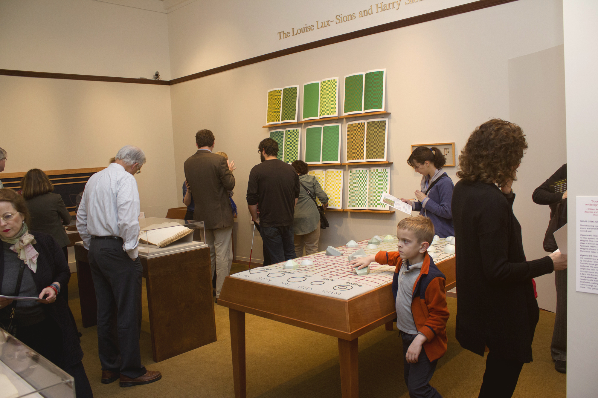 Picture shows several visitors, including a young boy, touching and viewing the installations in the center and east wall of the gallery. In the far left is a large book under a clear, mylar hood in a display case. In the left is Jayne’s Gift #5, a map representation of the travels the 19th–century blind surveyor John Metcalf. It is a vertically-positioned, large rectangular table composed of a linen top and a light brown wood base. A multi-color grid, outlines of geometric shapes, and green porcelain geometric shapes adorn the linen top. In the center background, on the back wall painted off-white, is Jayne’s Gift #4, a visual transmutation after the musical work of blind African American musician Thomas Wiggins. It is three horizontal and three vertical rows of prints in a geometric interplay of greens, browns and yellows. [end of description]