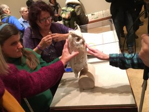 Picture shows exhibition visitors interacting with Teresa Jaynes’s Gift #2, a papier mâché owl on a wooden, sideways, cylindrical stand. In the right, the left hand of a man touches the front of the owl. The owl has yellow eyes and tan and white paper feathers. Part of the man’s right hand, which holds the top of a cane is also visible. In the left, a woman with long blond hair and wearing a green sweater touches the back of the owl. She stands between a women, with medium-length dark hair and wearing a purple zip-up cardigan, to her left, and the arm of another woman in a red sweater touching the back of the owl, to her right. The woman in purple, who also wears glasses, has her hands in fists and touching. Her knuckles touch in an explanatory gesture. Other gallery visitors walk and look at other parts of the exhibition in the background. [end of description]