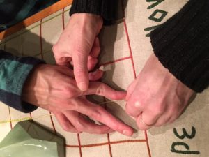 Picture shows the hands of exhibition visitors touching Teresa Jaynes’s Gift #5, a tactile map with an embroidered grid and green porcelain geometric shapes representing landforms on a linen background. In the lower right, the hands of a woman, the cuffs of her brown sweater visible, guide the hand of a man over the red threads of the embroidered grid. A part of one of the porcelain landforms is visible in the upper left of the image. [end of description]