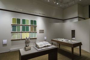 Picture shows the gallery, including four of Jaynes’s seven installations on view. In the center foreground is Jaynes’s Gift#1 on display on a flat, horizontally-angled rectangular table with a dark wood base. A white and brown paper owl rests on a cylindrical stand to the left of a large open book under a rectangular, clear acrylic hood. To the right of the display with the owl stands Jayne’s Gift #5, a map representation of the travels the 19th–century blind surveyor John Metcalf. It is a vertically-positioned, large rectangular table composed of a a linen top and a light brown wood base. A multi-color grid, outlines of geometric shapes, and green porcelain geometric shapes adorn the linen top. In the center background, on the back wall painted off-white, is Jayne’s Gift #4, a visual transmutation after the musical work of blind African American musician Thomas Wiggins. It is three horizontal and three vertical rows of prints in a geometric interplay of greens, browns and yellows. To the right of the prints on the wall is a small wooden frame in which brass musical notes are displayed. To the far right background is a view of Jaynes’s Gift #6, the scent mechanism the olfactometer, in a niche in an off-white curved wall. The floor of the room is covered with a tan colored carpet. [end of description]