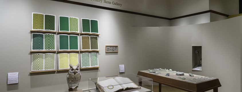 Picture shows the gallery, including four of Jaynes’s seven installations on view. In the center foreground is Jaynes’s Gift#1 on display on a flat, horizontally-angled rectangular table with a dark wood base. A white and brown paper owl rests on a cylindrical stand to the left of a large open book under a rectangular, clear acrylic hood. To the right of the display with the owl stands Jayne’s Gift #5, a map representation of the travels the 19th–century blind surveyor John Metcalf. It is a vertically-positioned, large rectangular table composed of a a linen top and a light brown wood base. A multi-color grid, outlines of geometric shapes, and green porcelain geometric shapes adorn the linen top. In the center background, on the back wall painted off-white, is Jayne’s Gift #4, a visual transmutation after the musical work of blind African American musician Thomas Wiggins. It is three horizontal and three vertical rows of prints in a geometric interplay of greens, browns and yellows. To the right of the prints on the wall is a small wooden frame in which brass musical notes are displayed. To the far right background is a view of Jaynes’s Gift #6, the scent mechanism the olfactometer, in a niche in an off-white curved wall. The floor of the room is covered with a tan colored carpet. [end of description]
