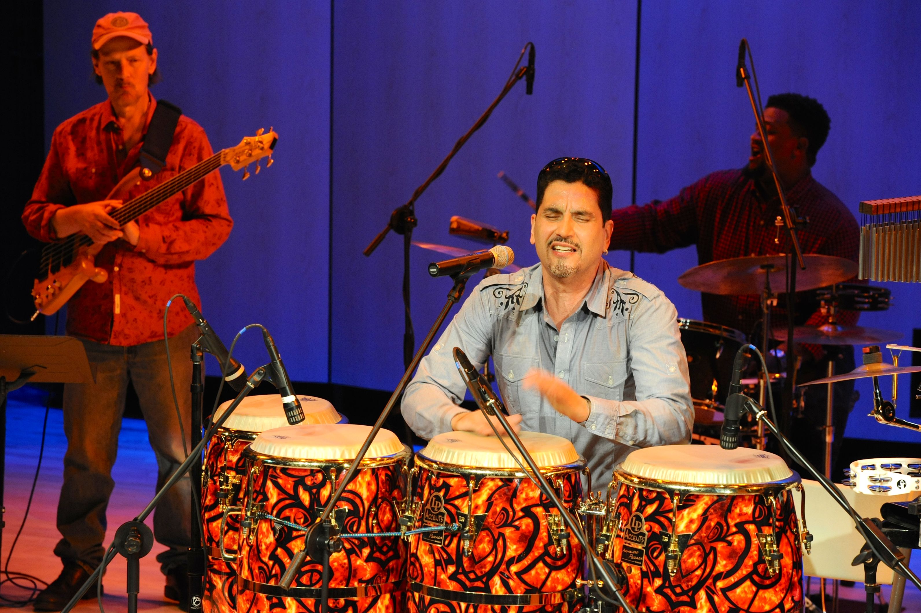 Picture shows Pablo Batista at his four-piece drum set during the middle of a concert. Batista has his eyes closed and his hands flutter above the drum in front of him. He wears a blue long sleeve shirt with dark blue ornamental details at the shoulders. A fellow musician with a guitar strapped over his shoulder stands to Batista’s right. Another drummer, seated at his drum set, plays behind Batista. [end of description]