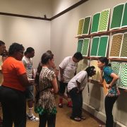 Photo shows a large group of individuals gathered in front of Teresa Jaynes’s Gift #4 of nine green, yellow, and brown screen prints. The screen prints depict grid patterns. In the right of the image, a young woman attired in black pants and a short-sleeved teal blouse with a flower pattern on the edge, slightly hunches over as if listening to something. Her right hand is to her ear and her left hand holds a large envelope to her waist. To her left, are two women. They lean and bend over in listening stances. In the left of the image, is a group of men and women in summer clothes. They look toward the women standing in front of the screen prints. The screen prints are hung above a short white-colored floor riser. [end of description]
